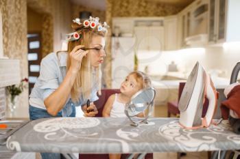 Housewife applying makeup at the ironing board, little baby looks at her. Woman with child at home together