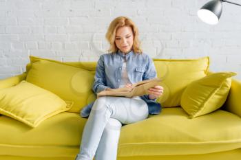 Young woman reading a book on cozy yellow couch, living room in white tones on background. Attractive female person with magazine sitting on sofa