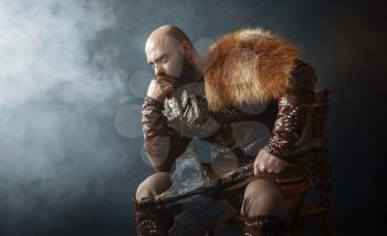 Thoughtful viking with axe dressed in traditional nordic clothes sitting on chair, barbarian image. Ancient warrior in smoke on dark background