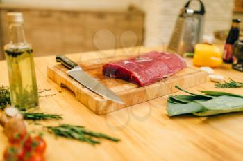 Raw meat tenderloin on wooden board closeup, nobody. Uncooked beef, vegetables, seasonings and spices with herbs on countertop, kitchen interior on background