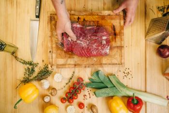 Male person seasoning piece of raw meat on wooden table, top view, kitchen interior on background. Chef cooking tenderloin with vegetables, spices and herbs