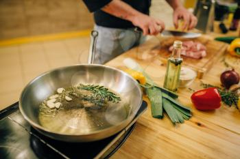 Frying pan with herbs and seasonings closeup, male person cuts meat. Man preparing boiled pork on table electric stove. Chef cooking tenderloin with vegetables on countertop
