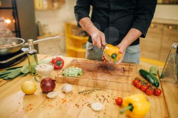 Male chef with knife cuts yellow pepper on wooden board, top view. Man cutting vegetables, fresh salad cooking, kitchen interior on background