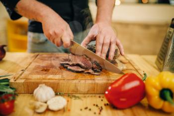 Male chef hands with knife cuts roasted meat on slices, top view. Man preparing beef with vegetables