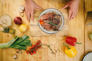 Male chef hands and roasted meat slices in a plate, top view, wooden countertop on background. Man preparing beef with vegetables, a gourmet dish