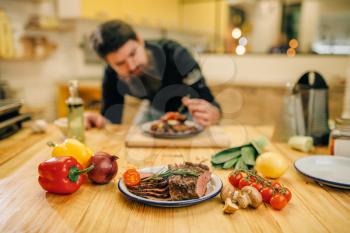 Male chef decorates with herbs a meat dish for gourmets, kitchen on background. Man preparing beef with vegetables on countertop