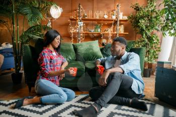 Black couple sitting on the floor and drinks coffee against a couch at home. Happy african love couple leisures in their house, cheerful family relaxing together in the morning