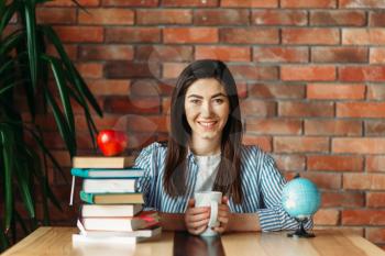 Female university student sitting at the table with textbooks and apple on the top, brick wall on background, knowledge concept. Young woman with books