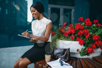 Business woman with notepad sitting on the bench during the break in front of office building. Smiling black businesswoman in white blouse works outdoors
