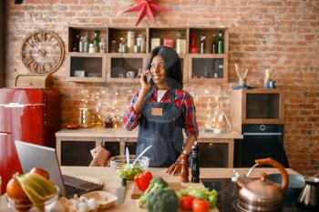 Black woman in apron talking by mobile phone on the kitchen. African female person preparing vegetable salad at home, ethnic girl cooking healthy breakfast