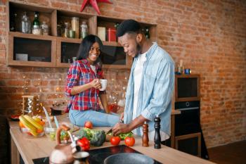 Black man cooking breakfast on the kitchen, wife drinks coffee. African couple preparing vegetable salad at home. Healthy vegetarian lifestyle