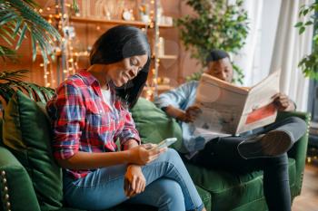 Black man reading newspaper, his woman using mobile phone on couch, relaxation at home. Happy african love couple leisures in their house