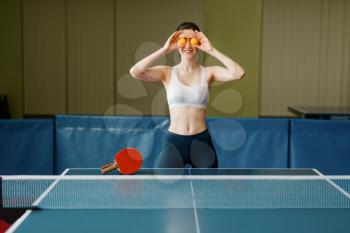 Young woman puts ping pong balls to her eyes indoors. Female person in sportswear, training in table-tennis club