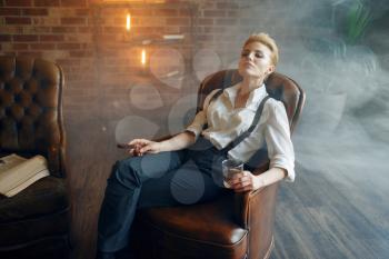 Woman in shirt and trousers sitting in leather chair with whiskey and cigar, retro fashion, gangster style