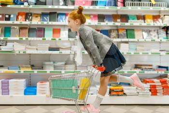 Little schoolgirl with a cart at the shelf, shopping in stationery store. Female child buying office supplies in shop, schoolchild in supermarket