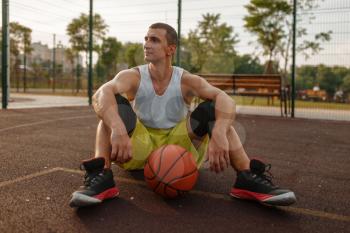 Basketball player sitting on the ground on outdoor court. Male athlete in sportswear resting after streetball training