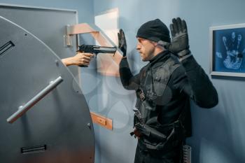 Bank robbery, male robber in black uniform standing at the vault door with his hands up. Criminal profession, theft concept