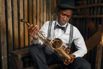 Black jazz musician sitting with saxophone, wooden fench background. Black jazzman in hat poses with instrument