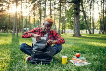 Black student with backpack sitting on the grass in summer park. A teenager studying outdoors and having lunch