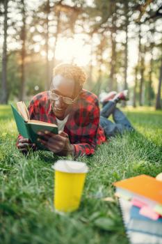 Black student in headphones lying on the grass and reading a book in summer park. A teenager studyingand leisures outdoors