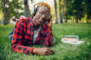 Black student in headphones resting on the grass in summer park. A teenager studying and leisures outdoors
