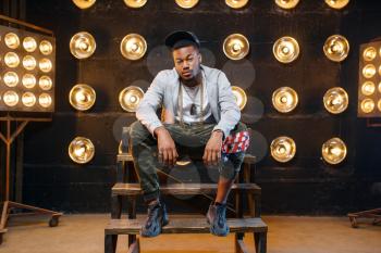 Black rapper in cap poses, perfomance on stage with spotlights on background. Rap performer on scene with lights, underground music