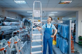 Male worker in uniform holds new aluminum stepladders in tool store. Department with ladders, choice of equipment in hardware shop, instrument supermarket