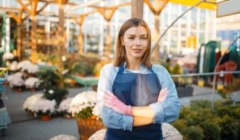 Female gardener poses in shop for gardening, saleswoman. Woman sells plants in florist store, seller in apron and gloves