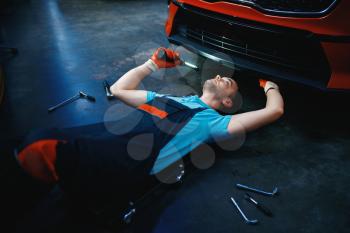 Worker in uniform lying under the vehicle, car service station. Automobile checking and inspection, professional diagnostics and repair