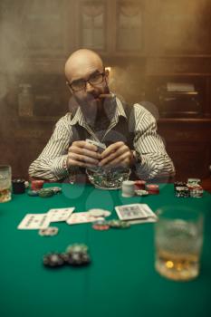 Bearded poker player with cigar, casino. Games of chance addiction. Man leisures in gambling house. Cards, chips and whiskey on gaming table