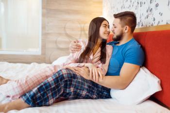 Cheerful love couple in pajamas hugs in bed at home, good morning. Harmonious relationship in young family. Man and woman resting together in their house, carefree weekend