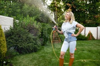 Smiling woman with hose watering trees in the garden. Female gardener takes care of plants outdoor, gardening hobby, florist lifestyle and leisure