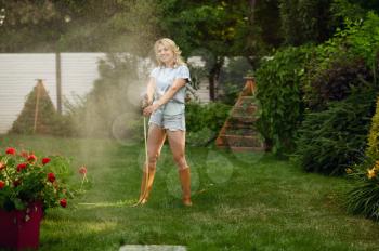 Attractive female gardener with hose watering plants in the garden. Woman takes care of flowers outdoor, gardening hobby, florist lifestyle and leisure
