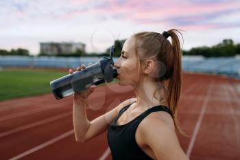 Female runner in sportswear drinks water, training on stadium. Woman doing stretching exercise before running on outdoor arena