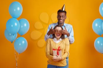 Happy man in cap congratulates his woman with gift box, yellow background. Pretty love couple, event or birthday celebration, balloons decoration