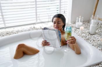 Sexy woman with glass reads a magazine in bath with milk. Female person in bathtub, beauty and health care in spa, wellness treathment in bathroom, pebbles and candles on background