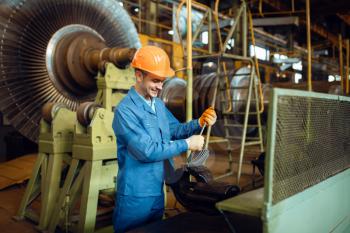 Engineer in uniform and helmet works with turbine detail on factory, impeller with vanes on background. Industrial production, metalwork engineering, power machines manufacturing