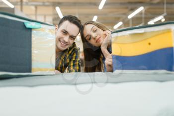 Young love couple choosing mattress for the bed in furniture store showroom. Man and woman looking samples for bedroom in shop, husband and wife buys goods for modern home interior