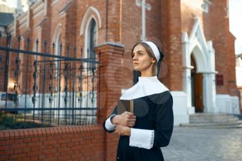 Young nun in a cassock holds a book, church on background. The sister is preparing for prayer in the monastery, religion and faith, religious people