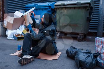 Dirty drunk beggar lies in garbage at the trashcan on city street. Poverty is a social problem, homelessness and loneliness, alcoholism and drunk addiction, urban lonely