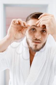 Man in bathrobe removes eyebrow hair in bathroom, routine morning hygiene. Male person at the sink performs skin and body treatment procedures