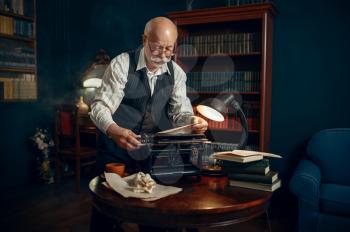 Elderly writer inserts paper into the vintage typewriter in his home office. Old man writes literature novel in room with smoke, inspiration, coffee and crumpled sheets on the table