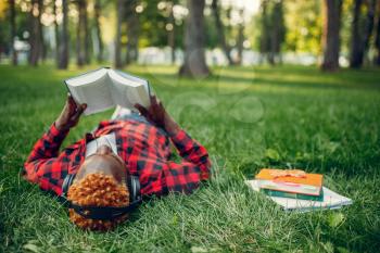 Black student in headphones resting on the grass in summer park, top view. A teenager studying and leisures outdoors