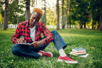 Black student in glasses writing in notebook on the grass in summer park. A teenager studying outdoors and having lunch