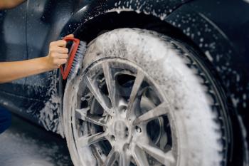 Female washer with brush in hand cleans wheel in foam, car wash. Woman washes vehicle, carwash station, car wash business