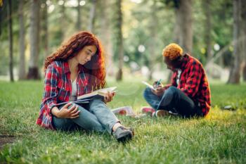 Students reading book on the grass in summer park. Male and female teenagers studying outdoors and having lunch