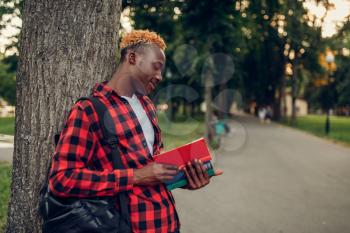 Black student with book and backpack standing near the tree in summer park. A teenager studying outdoors and having lunch