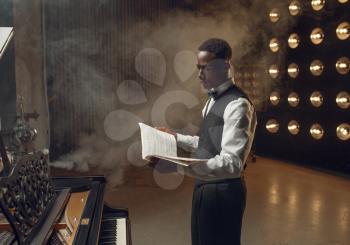 Ebony pianist with music notebook in his hands on the stage with spotlights on background. Negro performer poses at musical instrument before concert