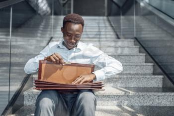 Negro businessman with briefcase sitting on stairs, mall. Successful business person, black man in formal wear, shopping center