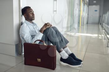 Ebony businessman with briefcase sitting on the floor in office hallway. Tired business person relax in corridor, black man in formal wear